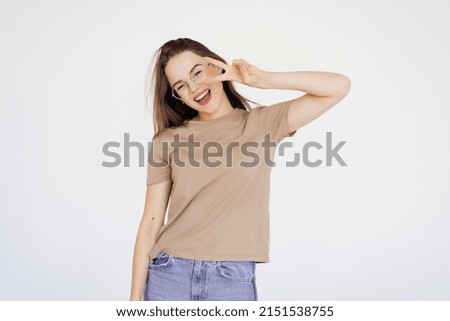 Cheerful young woman having fun, showing v-sign over eye and winking, smiling sassy at camera, being in good and positive mood, standing over white background