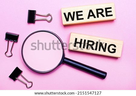 On a pink background, a magnifier, black paper clips and wooden blocks with the text WE ARE HIRING. Business concept