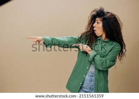 LOOK AT THIS CONCEPT. Overjoyed excited curly Latin female in casual things point finger aside posing isolated over pastel beige background look aside show copy space Mockup Banner. Good offer