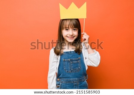Portrait of a cute brunette little girl holding a paper crown over her head playing princess with a smile Royalty-Free Stock Photo #2151585903