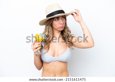 Young woman holding cocktail and wearing a bikini isolated on white background having doubts and with confuse face expression