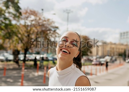 impressive girl with bright holiday make up is laughing outdoor in sunlight with happy smile on city background
