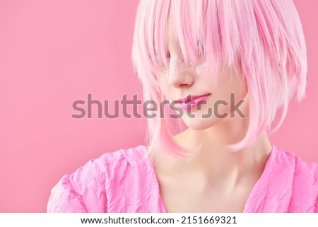 Asian style, anime. Portrait of a pretty teen girl with pink lips and glitter freckles posing in pink wig and dress on a pink background. Copy space.