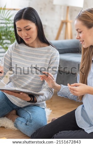 Two young girls learning foreign languages together at home sitting on the floor. College life, students learn. Concept of diverse friends studying together, preparation for exam. Startup, millennial