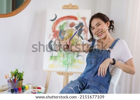 Creative of art concept, Young asian woman put paintbrush in mouth with artwork on canvas in studio.