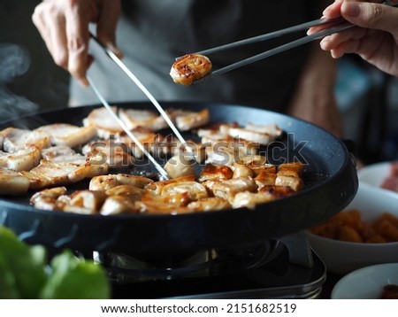 Female hand using silver chopsticks to eat Korean grilled pork large intestine, another person holding kitchen tongs to cook grilled pork belly or Samgyeopsal (Korean name) in BBQ non-stick grill pan. Royalty-Free Stock Photo #2151682519
