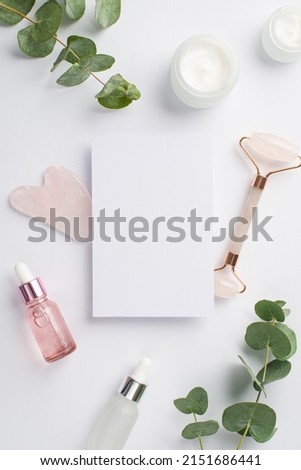 Skincare concept. Top view vertical photo of paper sheet rose quartz roller gua sha glass bottles with cosmetics and eucalyptus on isolated white background with blank space