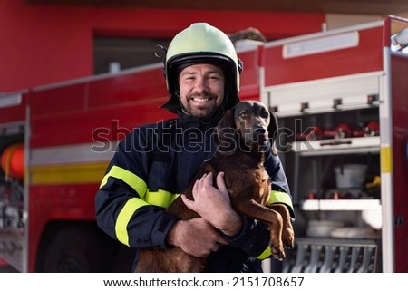 Happy firefighter man holding dog and looking at camera with fire truck in background