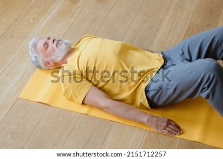 a man is lying on the floor in the shavasana pose and meditating. does yoga and fitness in the gym on a yellow mat. senior yoga at home or in the gym for training. a fifty-year-old man with Royalty-Free Stock Photo #2151712257