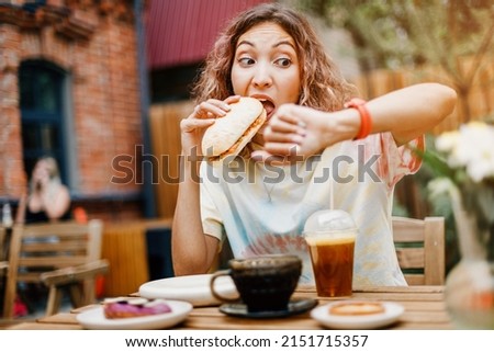 A girl in a hurry bites a burger on the run in a fast food cafe and looks at the smart watch. Late for a meeting and punctuality and digestive problems Royalty-Free Stock Photo #2151715357