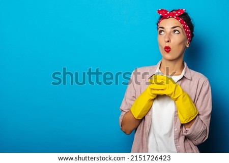 Surprised young woman in a bandage for hair, in gloves for cleaning looks up on a blue background