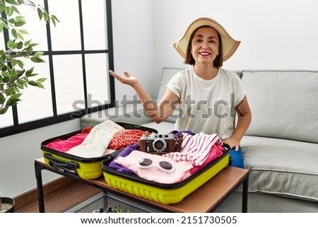 Beautiful middle age hispanic woman packing summer clothes in suitcase smiling cheerful presenting and pointing with palm of hand looking at the camera. 