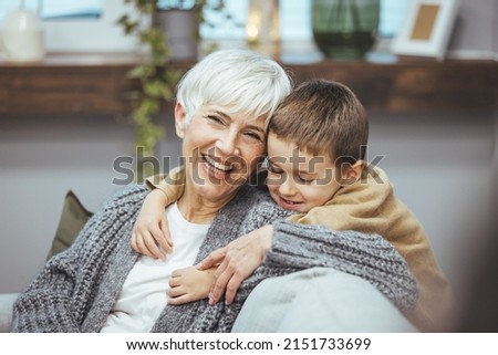 Grandma and grandson spend time together watching a funny video or cartoon on the phone, laughing while grandson hugs her. They enjoy their modern home.