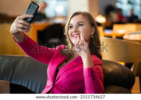 Photo of happy seductive woman winking and gesturing peace sign while taking selfie in a cafe