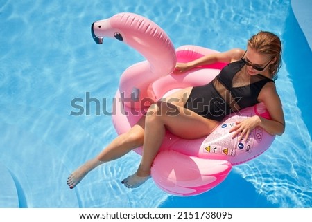 Hot slim woman relaxing on inflatable pink flamingo float mattress in bikini at swimming pool. Attractive fit girl in swimwear lies in the sun on floaty. Pretty female chilling on tropical vacation.