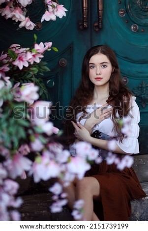 Beautiful long haired girl in medieval dress sitting on steps in front of blue door surrounded by magnolia flowers