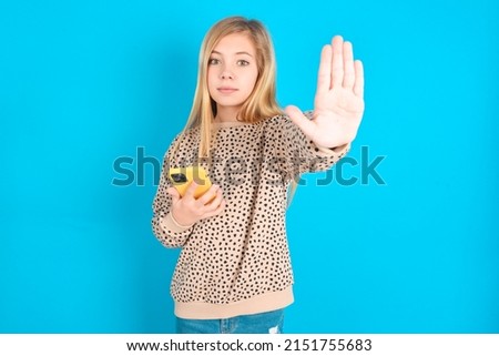 little caucasian kid girl wearing animal print sweater over blue background using and texting with smartphone with open hand doing stop sign with serious and confident expression, defense gesture