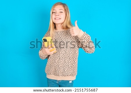 Portrait of little caucasian kid girl wearing animal print sweater over blue background using and texting with smartphone  happy with big smile doing ok sign, thumb up with fingers, excellent sign