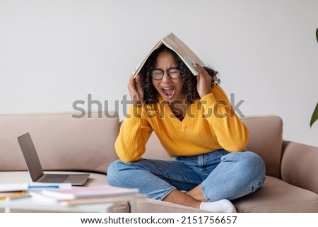 Young black woman shouting with book on her head near laptop computer at home, copy space. Emotional African American female having nervous breakdown because of too much online work or studies Royalty-Free Stock Photo #2151756657
