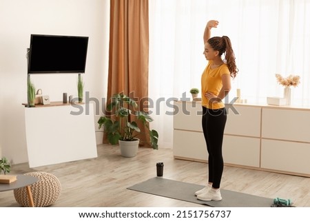 Sporty African American Woman Stretching Hands During Workout Watching TV With Empty Screen Standing On Gymnastic Mat Exercising At Home. Fitness Lifestyle Concept. Side View