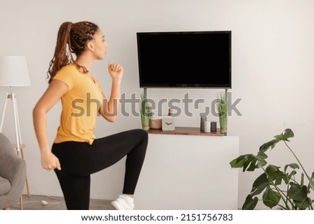 Sporty Woman Doing Knee To Elbow Leg Exercise During Workout Looking At Blank TV Screen At Home. Sporty Lifestyle And Domestic Fitness Training. Side View, Selective Focus
