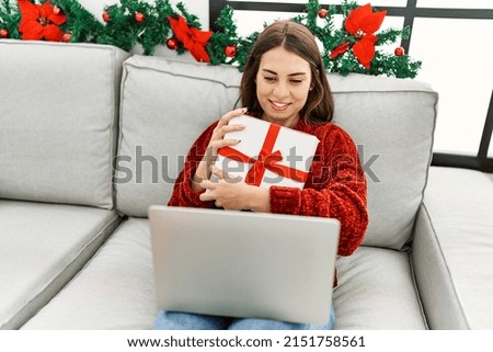 Young hispanic girl using laptop holding gift sitting on the sofa by christmas decor at home.