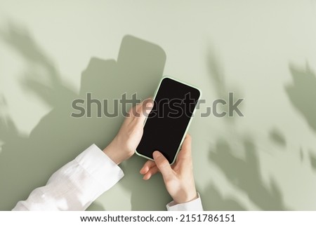 Mock up phone with black blank screen in female hands, white shirt sleeve, minimal flat lay on pastel green background. Summer lifestyle scene with smartphone, sunlight shadows, copy space, top view