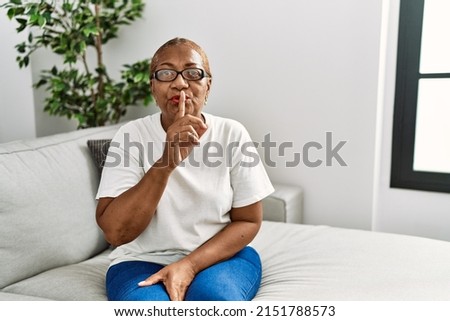 Mature hispanic woman sitting on the sofa at home asking to be quiet with finger on lips. silence and secret concept. 
