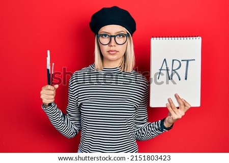 Beautiful blonde woman holding art notebook and brushes relaxed with serious expression on face. simple and natural looking at the camera. 