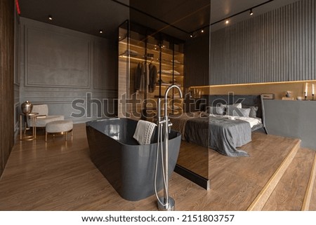 bedroom and freestanding bath behind a glass partition in a chic expensive interior of a luxury home with a dark modern design with wood trim and led light Royalty-Free Stock Photo #2151803757