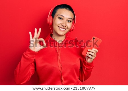 Beautiful hispanic woman with short hair using smartphone wearing headphones doing ok sign with fingers, smiling friendly gesturing excellent symbol 