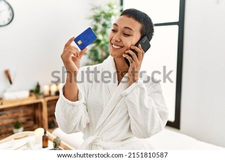 Young hispanic woman talking on the smartphone holding credit card at beauty center