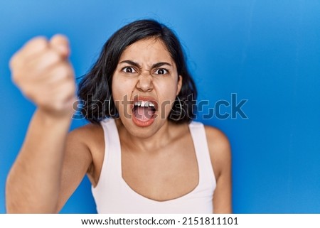 Young hispanic woman standing over blue background angry and mad raising fist frustrated and furious while shouting with anger. rage and aggressive concept.  Royalty-Free Stock Photo #2151811101