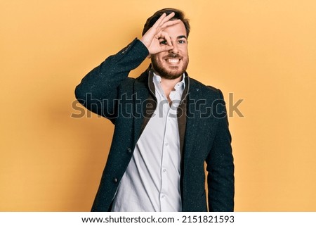 Handsome caucasian man with beard wearing elegant business jacket smiling happy doing ok sign with hand on eye looking through fingers 