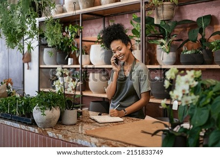 Smiling woman seller in botanical shop talking on smartphone. African american botanist writing client order in notebook during conversation on mobile phone in store. Young black small business owner. Royalty-Free Stock Photo #2151833749