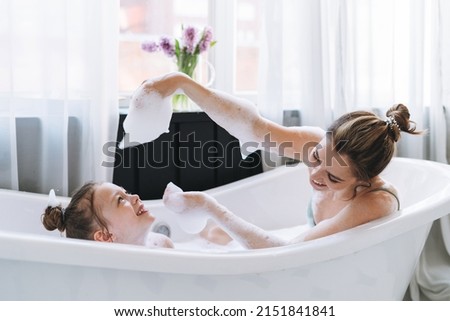Young mother woman with long hair with little tween girl daughter in pajamas having fun in the bath with foam at home