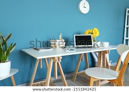 Workplace with modern laptop, books and vase with flowers near blue wall in room interior