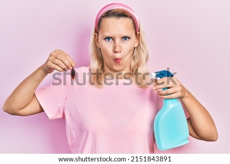 Beautiful caucasian blonde woman holding cockroach and pesticide sprayer making fish face with mouth and squinting eyes, crazy and comical. 