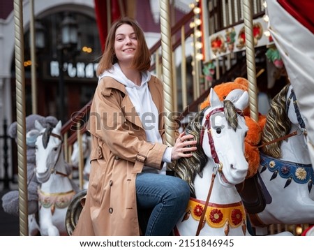 Beautiful woman in beautiful beige coat walk in autumn cloudy Moscow, enjoys and has fun on carousel. Street style. Travel to Russia concept. Royalty-Free Stock Photo #2151846945