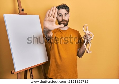 Caucasian man with beard standing by painter easel stand holding manikin with open hand doing stop sign with serious and confident expression, defense gesture 