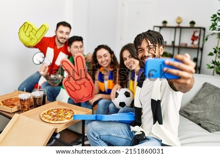 Group of young friends watching and supporting soccer match make selfie by the smartphone at home.