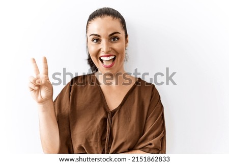 Young brunette woman standing over isolated background smiling with happy face winking at the camera doing victory sign. number two. 