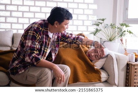 Man checking fever temperature of senior woman lying on bed. Mature husband resting at home feeling symptoms of the flu while wife checking fever by touching forehead.woman lying on the sofa blanket. Royalty-Free Stock Photo #2151865375