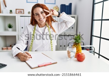 Young redhead woman nutritionist doctor at the clinic doing peace symbol with fingers over face, smiling cheerful showing victory 