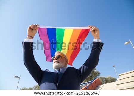 Mature gay man, executive, grey-haired, bearded, sunglasses, jacket and tie, waving the new lgbtiq+ pride flag in the wind under a blue sky. Mature gay man concept, lgbt, pride, equality, rights. Royalty-Free Stock Photo #2151866947
