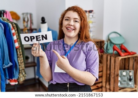 Young redhead woman holding banner with open text at retail shop smiling happy pointing with hand and finger 