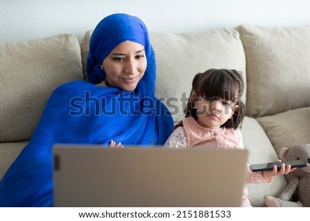 Young single Muslim mother with her young daughter using a laptop at home. Making video call, online entertainment, watching cartoons together and having fun.