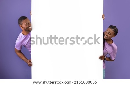Smiling young black people holding white vertical advertisement board, demonstrating free copy space for your text or design, positive guy and lady peeking out banner, purple violet background wall Royalty-Free Stock Photo #2151888055