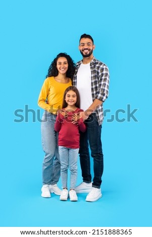 Cheerful Arabic Family Of Three Posing Together, Father And Mother Embracing Their Kid Daughter Standing On Blue Studio Background. Full Length, Vertical Shot