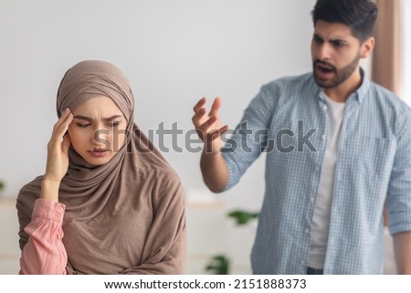 Domestic Violence. Mad Middle Eastern Husband Yelling At Scared Wife Sitting On Couch At Home. Selective Focus On Victimized Lady Wearing Hijab. Abuse And Aggression, Marital Problems Concept